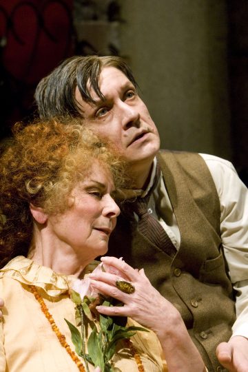 Kihara tukkainen nainen, jolla on keltainen mekko, pitelee vaaleanpunaista kukkaa ja nojaa rakastavasti ruskealiivinen ja kravattipukuinen mies, joka katsoo ylöspäin vakavalla ilmeellä.