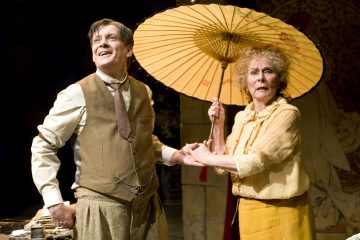 Vintagevaatteisiin pukeutunut mies hymyilee ja pitää kädestä kiinni keltaiseen mekkoon pukeutunutta huolestuneen näköistä naista, jolla on suuri koristeellinen aurinkovarjo päänsä yläpuolella. Molemmat seisovat lavalla dramaattisen valaistuksen alla.