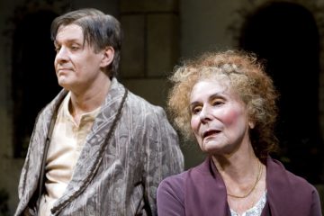 Vanhempi kiharatukkainen nainen ja tummahiuksinen mies seisovat lähekkäin lavalla, molemmilla on aikakauden puvut ja ilmeikkäät kasvot, jotka viittaavat teatteriesitykseen.