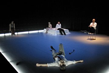 Neljä ihmistä istuu lavalla hämärässä valaistuksessa, ja yksi henkilö makaa etualalla lattialla levällään. Taustalla kaksi ihmistä jakaa sängyn, ja toinen istuu yksin tuolissa näyttämön oikealla puolella.