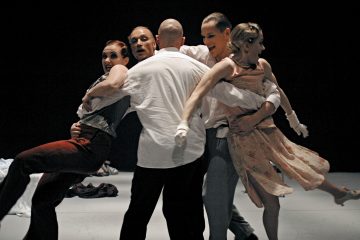 Neljä ihmistä tanssii tiiviisti yhdessä, ja kaksi miestä nostaa kahta naista sylissään. Kaikilla on yllään muodollinen tai puolimuodollinen asu, ja he näyttävät olevan liikkeessä tummaa taustaa vasten.