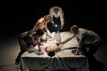 Neljä aikuista seisoo pimeässä huoneessa sängyllä valkoisen lakanan alla makaavan henkilön ympärillä ja valaisee häntä taskulampuilla samalla kun hän kurottautuu kohti, mikä luo dramaattisen, intensiivisen kohtauksen.