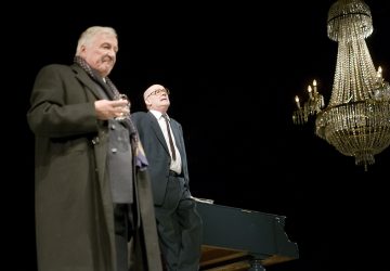 Lavalla on kaksi muodollisesti pukeutunutta iäkästä miestä, joista toinen seisoo etualalla lasia kädessään, kun taas toinen seisoo pianon ääressä. Heidän yläpuolellaan roikkuu suuri, koristeellinen kattokruunu tummaa taustaa vasten.