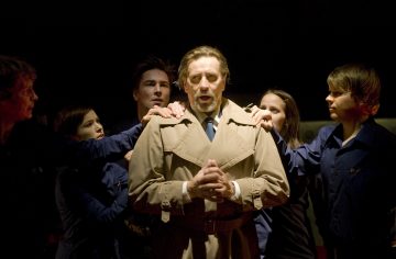 Ruskehtavaan juoksutakkiin pukeutunut mies seisoo käsi kädessä, silmät kiinni, ympärillään viisi tummansiniseen asuun pukeutunutta ihmistä, joista kukin laskee kätensä hänen olkapäilleen, dramaattisessa valaistuksessa.