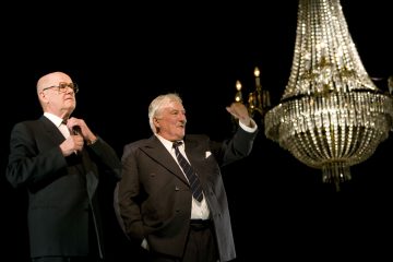 Kaksi vanhempaa pukumiestä seisoo suuren, koristeellisen kattokruunun alla pimeällä näyttämöllä. Toinen mies säätää silmälasejaan, kun taas toinen elehtii kädellään, mahdollisesti puhuu tai tervehtii yleisöä.