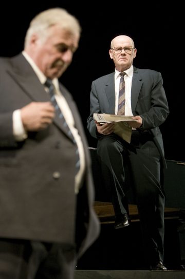 Lavalla on kaksi vanhempaa pukumiestä. Toinen seisoo etualalla epätarkasti ja säätää takkinsa, kun taas toinen seisoo taustalla tarkennettuna, paperit kädessään ja katsoo keskittyneesti eteenpäin.