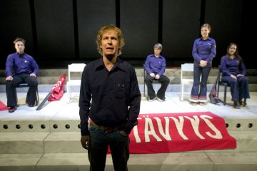 Mies seisoo näyttämön edessä ja puhuu tai laulaa, kun neljä ihmistä istuu hänen takanaan tuoleilla. Lavan portaiden yli on ripustettu punainen banderolli, jossa on valkoinen teksti. Tausta on tumma ja yksinkertainen.