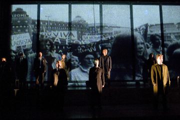 Lavalla seisoo joukko tummiin vaatteisiin pukeutuneita ihmisiä, ja heidän takanaan heijastetaan suuri mustavalkoinen kuva mielenosoituskohtauksesta, jossa on kylttejä ja väkijoukkoa. Tunnelma on dramaattinen ja vakava.