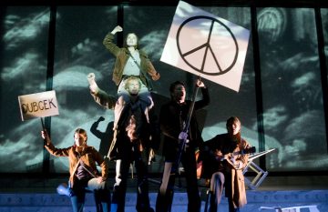 Lavalla viisi ihmistä pitää protestikylttejä, joista yhdessä on rauhansymboli ja toisessa lukee DUBCEK. Yksi henkilö kohoaa muiden yläpuolelle ja nostaa nyrkkiä. Taustalla on dramaattinen valaistus ja varjoisia pilviä.