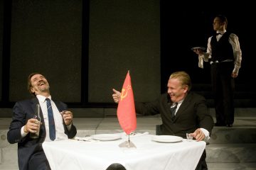 Kaksi pukumiestä nauraa ja juo pöydässä, jonka keskellä on Neuvostoliiton lippu, kun mustaliivinen tarjoilija seisoo taustalla tarjotin kädessään ja tarkkailee heitä.