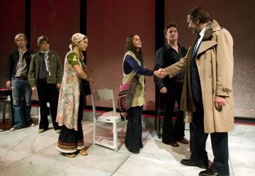 Kuusi ihmistä on lavasteissa. Liiviin pukeutunut nainen kättelee trenssitakkista miestä, ja neljä muuta katselee. Kaksi naista ja neljä miestä on pukeutunut retrotyylisiin vaatteisiin.