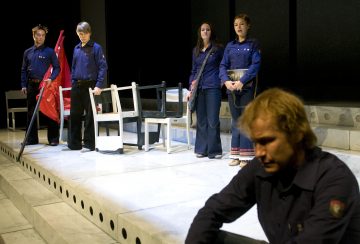 Viisi ihmistä on lavalla, jossa on valkoisia tuoleja; neljä seisoo taustalla, ja yksi istuu yksin etualalla ja katsoo mietteliäästi alaspäin. Kaikilla on tummansiniset paidat, ja kohtaus vaikuttaa dramaattiselta ja jännittyneeltä.