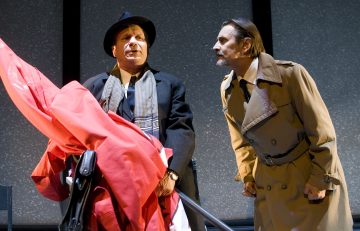 Kaksi miestä lavalla; toisella on hattu, huivi ja päällystakki, ja hänellä on punainen liina kädessään, ja toinen, jolla on ruskehtava juoksutakki, nojaa eteenpäin ja vaikuttaa intensiiviseltä. Taustalla on tavallinen, pilkullinen seinä.