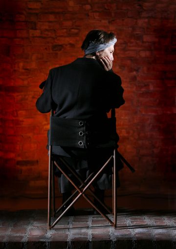 Henkilö, jolla on taaksepäin sidotut harmaat hiukset, istuu punatiiliseinää vasten taittotuolilla, nojaa päänsä yhteen käteen ja on pukeutunut tummaan takkiin takaapäin katsottuna.