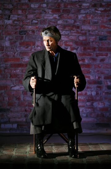 Tummaan takkiin ja huiviin pukeutunut iäkäs henkilö istuu tuolilla punatiiliseinää vasten, tarttuu käsinojiin ja katsoo sivulle dramaattisessa valaistuksessa.