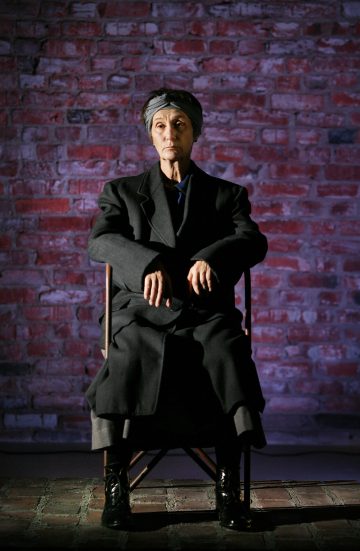 Tummaan takkiin ja huiviin pukeutunut vanhempi henkilö istuu tuolilla punatiiliseinää vasten, kädet polvillaan, ja katsoo mietteliäästi sivulle dramaattisessa valaistuksessa.