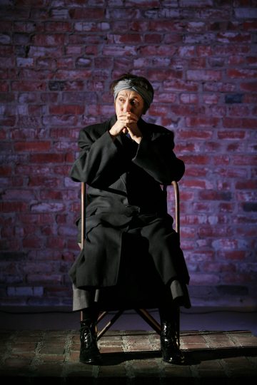 Tummiin vaatteisiin ja huiviin pukeutunut henkilö istuu tuolilla punaista tiiliseinää vasten ja näyttää mietteliäältä kädet suunsa lähellä ja jalat ristissä.