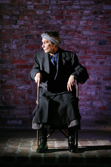 Henkilö, jolla on tumma puku, pitkä hame, saappaat ja päähine, istuu tuolilla punatiiliseinää vasten ja katsoo vasemmalle. Kohtaus on dramaattisesti valaistu.