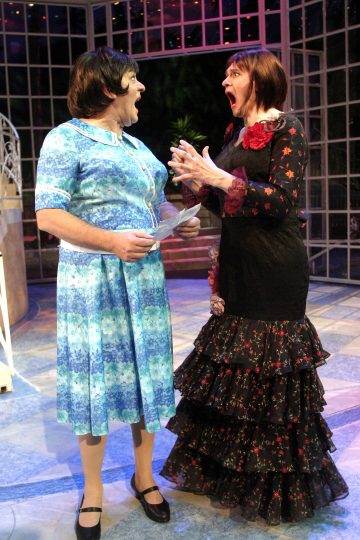 Lavalla on kaksi näyttelijää, toinen sinikuvioisessa mekossa ja toinen pitkässä mustassa röyhelömäisessä mekossa, jossa on punaisia kukkia, ja he ovat vastakkain hämmästyneillä ja ilmeikkäillä kasvoillaan kirkkaasti valaistussa lavastuksessa.