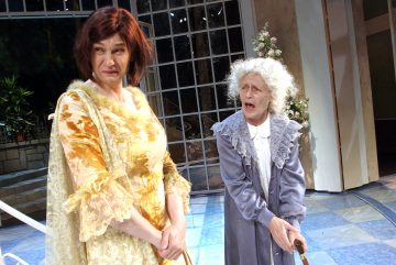 Lavalla esiintyy kaksi näyttelijää, joista toinen on pukeutunut keltaiseen mekkoon ja peruukkiin ja vaikuttaa skeptiseltä, kun taas toinen, harmaisiin vaatteisiin ja valkoiseen peruukkiin pukeutunut näyttelijä näyttää yllättyneeltä ja pitelee keppiä. Taustalla on suuri ikkuna ja kasveja.