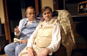 Kaksi vanhaan pukuun pukeutunutta miestä istuu lähekkäin junassa, toinen sinipukuinen mies pitelee sanomalehteä ja elehtii, toinen beige pukuinen ja liivi, molemmat katsovat keskittyneesti kameraan.