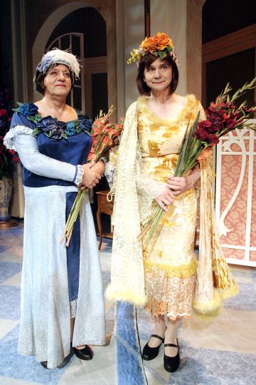 Kaksi värikkäisiin, vanhanaikaisiin mekkoihin ja kukkapäähineisiin pukeutunutta ihmistä seisoo sisätiloissa, kummallakin kukkakimppu kädessään, todennäköisesti teatteriympäristössä, jossa on koristeltu väliseinä ja kukkia taustalla.