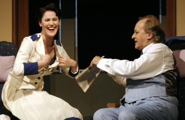 Valkoiseen laivastopukuun pukeutunut nainen nauraa istuessaan vaaleansiniseen pukuun pukeutuneen, paperia kädessään pitävän miehen vieressä.