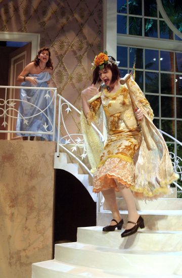 Kaksi naista esiintyy teatterilavalla. Toinen, jolla on kultainen mekko ja kukkainen päähine, seisoo keskellä portaita ja tekee leikkisän eleen. Toinen, siniseen mekkoon pukeutunut nainen seisoo hänen takanaan portaikossa ja reagoi vilkkaasti.