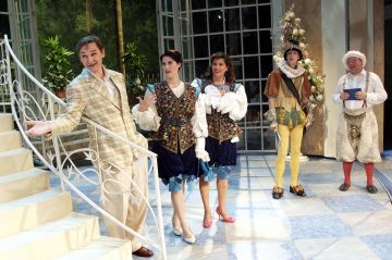 Viisi näyttelijää värikkäissä, taidokkaissa puvuissa esiintyy valoisalla, tyylikkäällä näyttämöllä, jossa on suuret ikkunat ja kasveja; yksi mies seisoo portailla elehtien, kun muut katselevat ja reagoivat ilmeikkäästi.