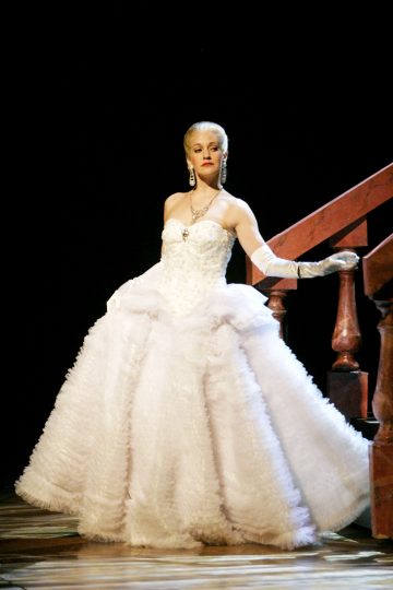 Nainen lumoavassa valkoisessa tanssiaispuvussa, pitkissä hanskoissa ja kimaltelevissa koruissa seisoo tyylikkäästi portaiden vieressä poseeraten tummaa taustaa vasten.