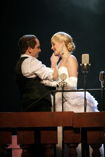 Pukumies koskettaa naisen leukaa, kun he seisovat lähekkäin mikrofonien takana lavalla. Naisella on valkoinen puku ja käsineet, ja molemmat näyttävät esiintyvän dramaattisessa kohtauksessa.