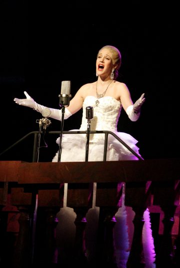 Lumoavaan valkoiseen pukuun ja hanskoihin pukeutunut nainen laulaa intohimoisesti lavalla oleviin vintage-mikrofoneihin, joita valaisee kirkas valokeilavalo tummaa taustaa vasten.