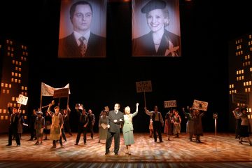 Näyttämökohtaus, jossa näyttelijöillä on mielenosoituskylttejä, kaksi ihmistä keskellä näyttämöä ja yläpuolella roikkuvat suuret miehen ja naisen muotokuvat. Taustalla on tyyliteltyjä kaupunkirakennuksia.