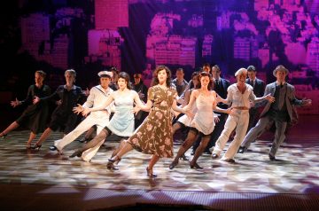 Ryhmä vintage-asuihin pukeutuneita esiintyjiä tanssii tarmokkaasti lavalla, jonka taustalla on violetisti valaistu kaupunkimaisema, keskellä ruskeaan mekkoon pukeutunut nainen.