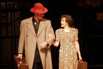 Pitkätakkinen ja punahattuinen mies pitelee matkalaukkua ja kättelee hymyilevää kuviomekkoon pukeutunutta naista, joka myös kantaa matkalaukkua, molemmat lavalla lämpimässä valaistuksessa.