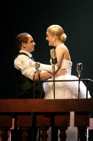 Kaksi virallisesti pukeutunutta näyttelijää, toinen liivissä ja toinen valkoisessa puvussa, seisovat lähekkäin ja syleilevät toisiaan lavalla vanhojen mikrofonien takana ja katsovat toisiaan dramaattisessa valaistuksessa.