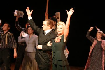 Kaksi näyttelijää seisoo edessä ja nostaa kätensä kumartaen, kun muut pukuun pukeutuneet näyttelijät juhlivat ja heiluttavat sanomalehtiä taustalla teatterin lavalla.