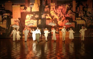 Ryhmä vintage-asuihin pukeutuneita esiintyjiä, joilla on sateenvarjot kädessään, tanssii rivissä lavalla, ja taustalla on kirkkaasti valaistu kaupungin horisontti auringonlaskun aikaan.