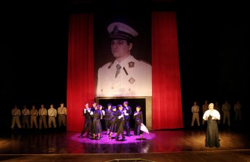Teatteriesitys, jossa näyttelijät ovat tummissa puvuissa ja lavalla on laulaja pitkässä puvussa. Heidän takanaan roikkuu suuri muotokuva valkoiseen sotilaspukuun pukeutuneesta miehestä, jota reunustavat punaiset verhot ja valkoisiin univormuihin pukeutuneet ihmiset.