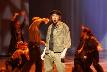 Che Guevara -tyyliseen asuun pukeutunut mies seisoo näyttämön keskellä, suu auki ikään kuin laulaisi tai puhuisi, ja hänen takanaan on useita ihmisiä, jotka elehtivät energisesti dramaattisessa valaistuksessa.