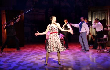 Kukkamekkoon pukeutunut nainen seisoo näyttämön keskellä ja laulaa kädet levällään, kun useat vintage-pukuiset ihmiset poseeraavat dramaattisesti hänen ympärillään teatterivalaistuksessa.