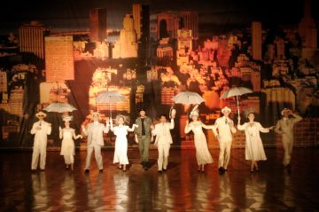 Joukko valkoisiin pukuihin pukeutuneita esiintyjiä, joilla on sateenvarjot kädessään, tanssii rivissä näyttämön poikki, ja heidän takanaan on lämpimillä valoilla valaistu kaupungin horisontti.