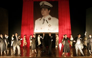 Vanhoihin juhlavaatteisiin pukeutuneet esiintyjät seisovat lavalla laulaen ja elehtien; heidän takanaan roikkuu suuri muotokuva valkoiseen sotilasunivormuun pukeutuneesta miehestä punaisia verhoja vasten.