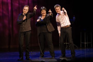 Lavalla on kolme ihmistä, joilla jokaisella on mikrofoni kädessään ja jotka hymyilevät ja osoittavat kohti yleisöä. He seisovat punaisen verhon edessä näyttämövalojen alla.