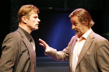 Kaksi pukuihin pukeutunutta miestä käy jännittävää keskustelua lavalla. Toinen mies katsoo vakavalla ilmeellä eteenpäin, kun taas toinen mies elehtii häntä kohti nostetulla kädellä ja näyttää selittävän tai väittelevän.