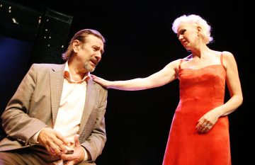 Nainen hihattomassa punaisessa mekossa laskee kätensä varovasti istuvan, vaaleaan puvuntakkiin pukeutuneen miehen olkapäälle, ja molemmat katsovat toisiaan dramaattisessa näyttämövalaistuksessa.