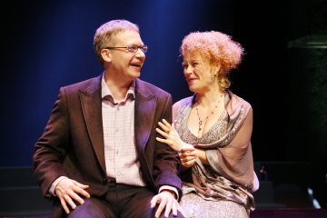 Mies ja nainen istuvat lähekkäin lavalla hymyillen ja nauraen kirkkaiden valojen alla. Miehellä on silmälasit ja puvun takki; naisella on hopeinen mekko ja huivi, ja hänellä on kiharat hiukset, ja hän pitelee hellästi miehen kättä.