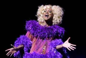 Henkilö, jolla on kiharat vaaleat hiukset, laulaa intohimoisesti yllään värikäs mekko, jossa on violetteja ja vaaleanpunaisia pyörteitä, ja dramaattinen violetti höyhenboa mustaa taustaa vasten.