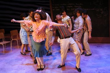 Ryhmä värikkäisiin pukuihin pukeutuneita näyttelijöitä tanssii ja hymyilee lavalla, ja edessä on nainen, joka ojentaa kätensä, ja mies, joka nojaa taaksepäin hänen vieressään. Taustalla on tuoleja ja liitutaulu, johon on kirjoitettu.