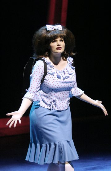 Lavalla olevalla naisella on suuri rusetti hiuksissaan, pilkkupusero, sininen hame, jossa on röyhelöinen helma, ja valkoiset käsineet. Hän elehtii avosylin ja näyttää ilmeikkäältä, ja häntä valaisee näyttämövalaistus tummaa taustaa vasten.